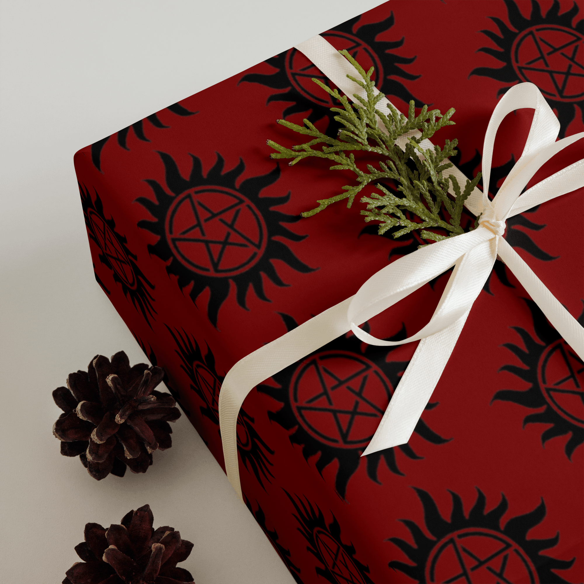 Square gift box wrapped in red paper with black circular symbol patterns, tied with white ribbon and accented with greenery and pinecones.