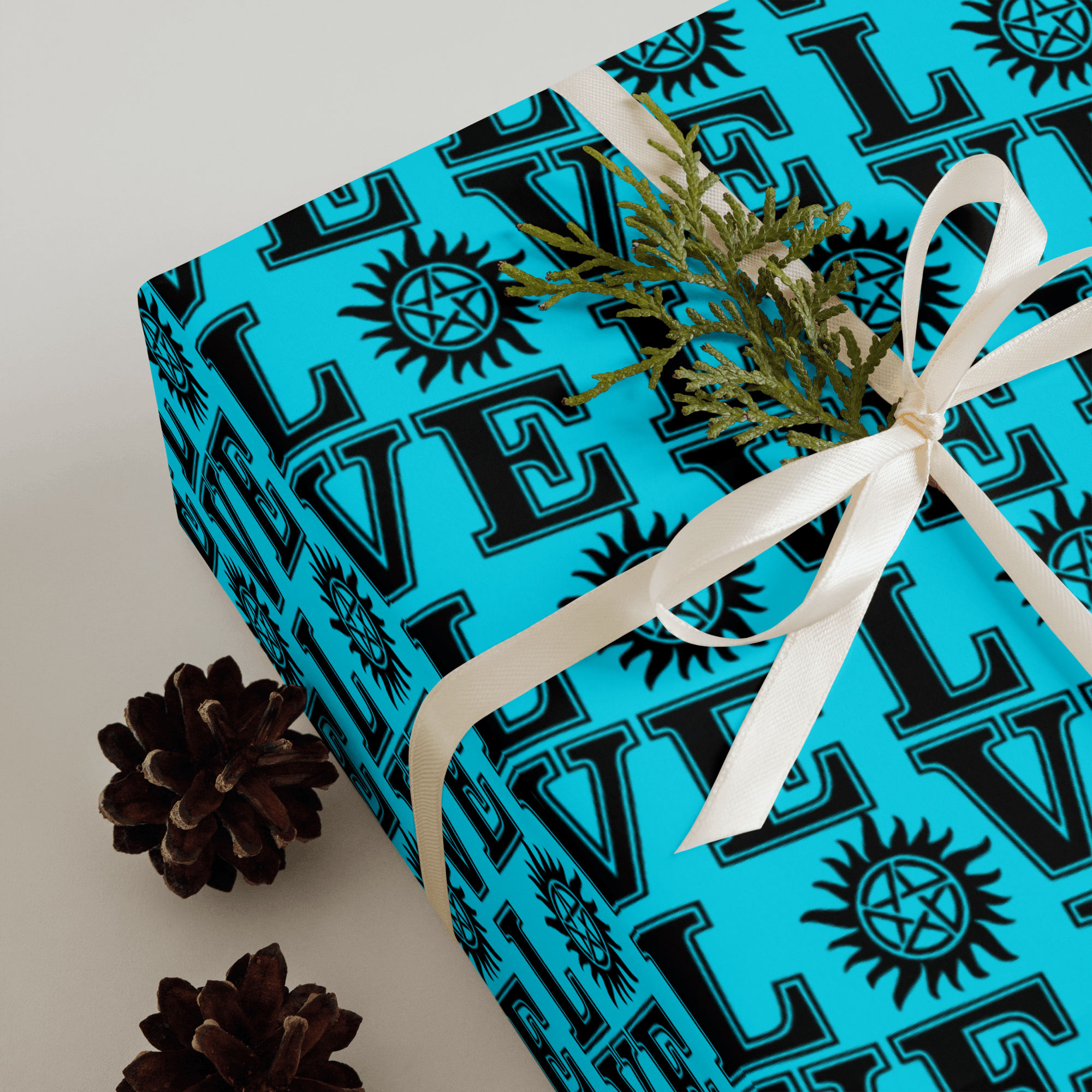 Square gift box wrapped in teal paper with bold repeating graphic symbols, tied with white ribbon and styled with pinecones.